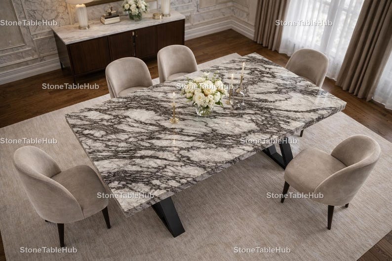 May include: A rectangular marble dining table with a black base, surrounded by six beige upholstered chairs. The table has a white and gray veined pattern. A vase of white flowers and candles adorn the table. The room has a neutral color palette.