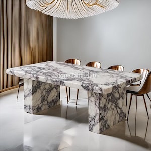 May include: A rectangular marble dining table with a white and purple veined pattern. The table has a thick, solid base and is surrounded by bronze-coloured chairs. The room features a large chandelier and a wooden wall panel.