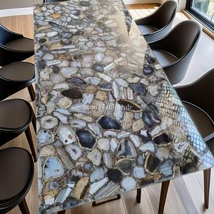 Op de afbeelding: Een rechthoekige eettafel met een mozaïek stenen blad in tinten grijs, blauw en bruin. De tafel is omringd door zwarte stoelen met houten poten. De tekst "StoneTablehub" is zichtbaar op het tafeloppervlak.