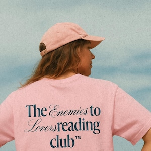 Puede incluir: Una camiseta rosa claro con el texto azul marino "The Enemies to Lovers reading club". Se lleva una gorra de béisbol rosa a juego. El fondo es azul claro.