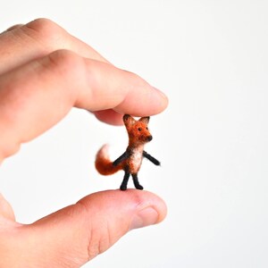 May include: A small felt fox figurine, held between fingers. The fox has a reddish-orange body, a white-tipped tail, and black accents on its legs and ears. The fox is approximately 2.5 cm tall.