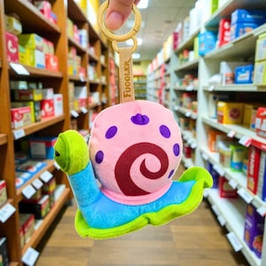May include: A plush snail keychain with a pink body, purple spots, and a maroon swirl design. The snail has a blue base and green accents. The keychain has a gold ring and a tan strap with the word "FUGGLER".