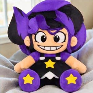 Pode incluir: Boneca de pelúcia com um personagem de desenho animado com cabelo roxo e preto, olhos grandes e um sorriso largo. A boneca usa uma roupa preta com detalhes de estrelas amarelas e detalhes roxos.