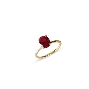 May include: A gold ring featuring a vibrant red oval-cut gemstone. The ring's band is a thin, polished gold, and the gemstone is securely held by four prongs. The design is simple yet elegant, suitable for various occasions.
