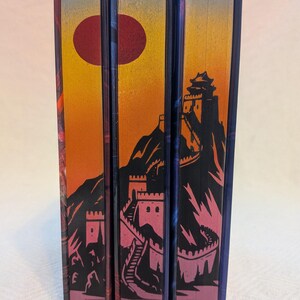 May include: A stack of three books with colorful, illustrated spines. The spines feature a sunset scene with a red sun, mountains, and a fortress. The color palette includes orange, pink, black, and blue.