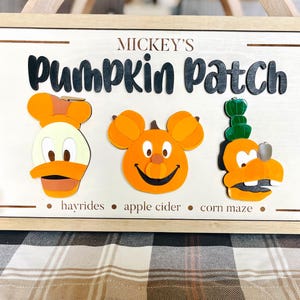 May include: Wooden sign with the text "Mickey's Pumpkin Patch" and three cartoon character pumpkins. The sign includes the phrases "hayrides", "apple cider", and "corn maze". The sign is framed in a light brown wood.