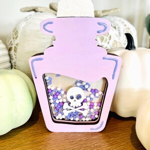 Könnte beinhalten: Eine hellviolette Holzbottichflasche mit einem Totenkopf- und gekreuzten Knochen-Design. Die Flasche ist mit kleinen Perlen in Weiß, Lila und Blau gefüllt. Die Flasche ist mit hellvioletter Farbe und einem weißen Verschluss verziert.