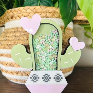 May include: A light green cactus-shaped decoration with a pink base and heart accents. The cactus has a clear center filled with small, colorful beads and confetti. The base features a geometric design.