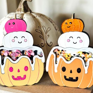 Op de afbeelding: Twee Halloween-pompoenversieringen. Elk heeft een lachende jack-o'-lantern-basis, een witte geest en een kleine pompoen bovenop. De bases zijn oranje en geel, met een transparant gedeelte gevuld met kleurrijke confetti.
