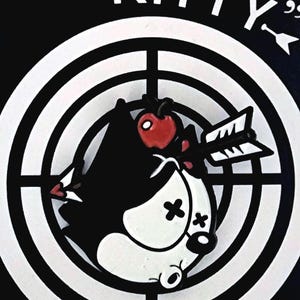 May include: A black and white graphic pin featuring a cartoon cat with an arrow through its head, set against a target. The text "POOR KITTY" is above the cat. The pin has a red apple on its head.