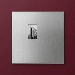 Puede incluir: Una placa de pared cuadrada de metal plateado cepillado con un recorte rectangular central. Dentro de este recorte hay una fotografía en blanco y negro de un edificio moderno con una fachada alta y angular y múltiples ventanas.