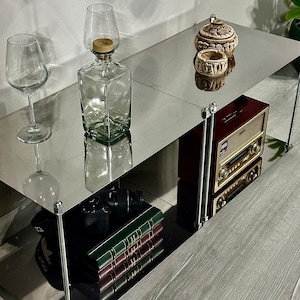 May include: A modern, mirrored shelf unit displaying decorative items. Includes a clear glass wine glass, a square glass bottle with a wooden stopper, a small decorative jar, a vintage radio, and a stack of books with green and burgundy covers.