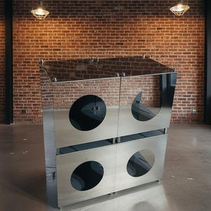 May include: A modern, stainless steel cabinet with a mirrored top and four circular cutouts. The cabinet has a unique design and is set against a brick wall background. The cabinet is likely used for storage.