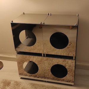 May include: A mirrored cabinet with four square compartments, each featuring a large, circular opening. The cabinet is made of a reflective, silver material and sits on a light-colored rug. The design is modern and minimalist.