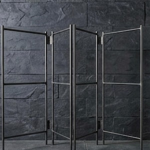 以下が含まれることがあります： 暗い金属製の4パネルのパーテーション。各パネルは水平バーのある長方形のフレームです。パネルはヒンジで接続されており、折りたたむことができます。パーテーションは、暗く質感のある壁に立てかけられています。