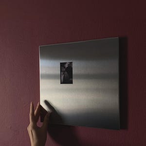 Könnte beinhalten: Eine rechteckige Magnettafel aus gebürstetem silberfarbenem Metall mit einer glatten, reflektierenden Oberfläche. Ein kleines, vertikales Schwarz-Weiß-Foto ist in der Mitte der Metallplatte befestigt und wird durch magnetische Kraft gehalten.