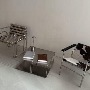May include: A silver metal chair with horizontal tubular bars, a square two-tiered metal coffee table with a small lamp and two books, and a chair with a chrome frame, black armrests, and a brown and white cowhide seat and backrest.