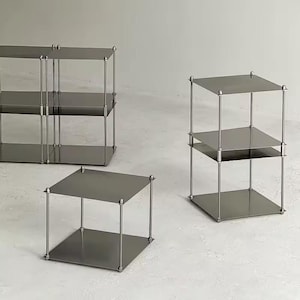 Puede incluir: Tres mesas auxiliares modernas, de color gris metálico, con un diseño minimalista. Cada mesa tiene una parte superior cuadrada y dos estantes, soportados por finos postes verticales con remates esféricos decorativos. Las mesas están dispuestas sobre una superficie clara.
