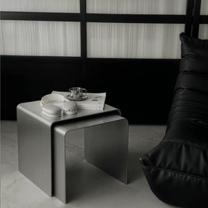 May include: A set of two nested tables made of brushed silver metal with a smooth, curved U-shaped design. The tables are stacked together, with a white book, a small metallic cup, and two white rounded objects resting on the top surface.