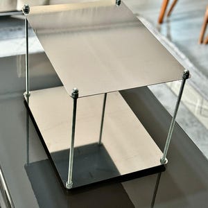 May include: A two-tiered, silver-coloured metal side table. The table has a rectangular top and bottom, connected by four vertical metal rods secured with nuts and bolts. The table is approximately 46 cm long, 30 cm wide, and 41 cm tall.