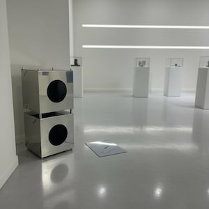 May include: A stainless steel cube-shaped display case with two large circular openings. The case is stacked on another identical case. The cases are in a gallery setting with white walls and pedestals.