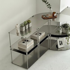 May include: A multi-tiered modular shelving unit constructed from brushed stainless steel with thin vertical support rods. The unit features rectangular and semi-circular shelves holding books, small potted succulents, a glass vase, and a picture frame.