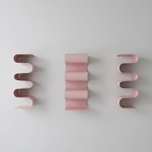 May include: Three pink metal wall shelves with a wavy, ribbon-like design. The shelves are mounted on a white wall, with one shelf in the center and two on either side. The shelves are a soft, pastel pink color.