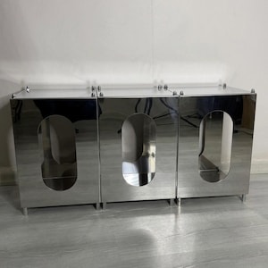 May include: A mirrored cabinet with three doors and oval cutouts. The cabinet is made of a reflective, silver-toned material and has small, silver feet. The top of the cabinet has small, decorative silver accents.