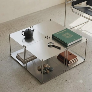 May include: A square, two-tiered metal coffee table with a brushed finish and thin vertical supports. On the top surface sits a black teapot, a green book titled Aesthetic Modules, and a magnifying glass. The lower shelf holds folded fabric and stones.
