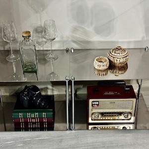 Puede incluir: Una exhibición de artículos vintage en un estante espejado. Incluye dos copas de vino, una botella de vidrio transparente con tapón de madera, un pequeño tarro decorativo, una radio vintage roja y dorada, una cámara y una pila de libros.