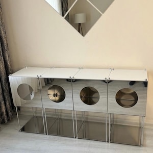 May include: A modern, mirrored cabinet with four compartments, each featuring a circular opening. The cabinet has a white top and sits on slender, silver legs. A decorative mirror is mounted on the wall above the cabinet.