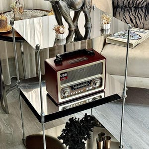 May include: A three-tiered mirrored side table with chrome supports. The top shelf holds a silver elephant statue and small white vases. The middle shelf displays a vintage red radio. The bottom shelf features a black coral decoration and small gold cups.