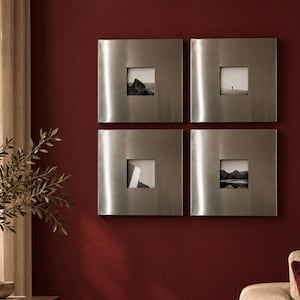 May include: A set of four square wall frames with a brushed silver metallic finish. Each frame features a wide, smooth border and a central square cutout displaying a small black and white photograph of a landscape or abstract scene.