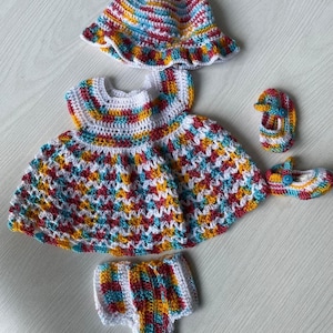May include: A colorful crochet baby outfit set, including a dress, hat, bloomers, and two pairs of shoes. The set features a mix of white, yellow, red, and blue yarn. The dress has a round neckline and a flared skirt.