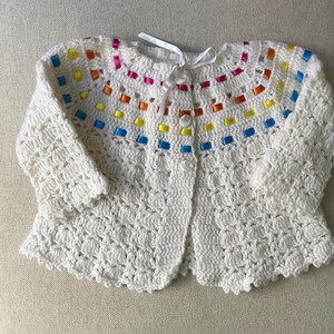 May include: A white, crocheted baby cardigan with colorful ribbon accents. The sweater features a scalloped hem, button closure, and a ribbon tie at the neckline. The colorful ribbon is in shades of pink, orange, yellow, and blue.