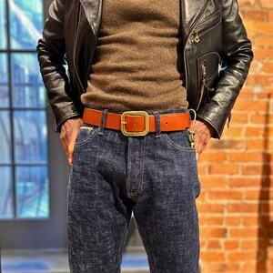 May include: A brown leather belt with a gold-colored square buckle is worn with blue denim jeans. The belt is approximately 1.5 inches wide. The person is wearing a brown sweater and a black leather jacket.