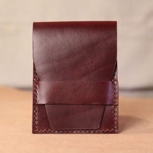 May include: A vertical, rectangular dark burgundy leather card holder featuring a smooth front flap and a horizontal strap across the lower section. The edges are finished with visible, thick tonal stitching, creating a structured, compact design.