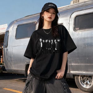 May include: A black short-sleeved t-shirt featuring a monochromatic graphic print of a group of seven individuals standing in a row. The shirt has a crew neckline and a loose, relaxed fit. The graphic is positioned centrally on the chest of the garment.