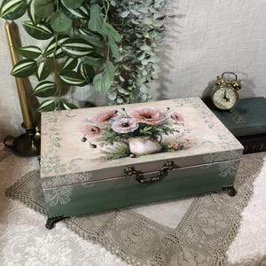 Puede incluir: Caja de madera decorativa con un diseño floral en la tapa. La caja está pintada en tonos verdes y crema, con un asa y patas de latón. La tapa presenta un ramo de flores rosas y blancas en un jarrón blanco.
