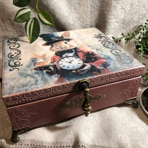 Peut inclure: Coffret décoratif en bois avec couvercle à charnière, orné d'une illustration fantaisiste d'un lapin en gilet et haut-de-forme tenant une montre de poche et une assiette de gâteau. Le coffret a une quincaillerie et des pieds en métal ouvragé.
