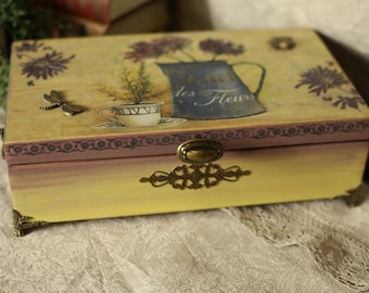 Puede incluir: Una caja decorativa de madera con un diseño floral. La caja presenta una pintura degradada amarilla y rosa, un cierre de metal y patas de esquina decorativas. La tapa está adornada con una jarra azul, flores y el texto "des Fleurs".