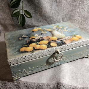 May include: A rectangular wooden box with a hinged lid, painted in shades of blue and adorned with a lemon still life. The lid features a painting of lemons, a pitcher, and books. The box has ornate carvings and a decorative handle.