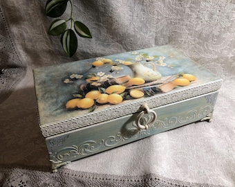 Puede incluir: Una caja rectangular de madera con tapa con bisagras, pintada en tonos azules y adornada con un bodegón de limones. La tapa presenta una pintura de limones, una jarra y libros. La caja tiene tallas ornamentadas y un asa decorativa.