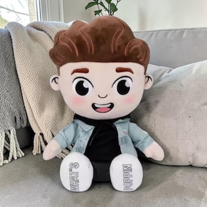Puede incluir: Muñeco de peluche de un niño de dibujos animados con cabello castaño, que lleva una chaqueta vaquera azul claro y una camisa negra. El muñeco tiene ojos grandes y una cara sonriente. Los pies del muñeco dicen "WHAT'S POPPIN".