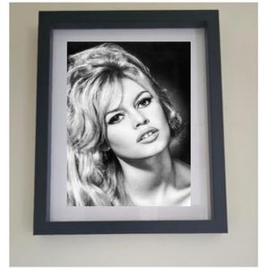 May include: Black and white framed portrait of a woman with blonde hair. The portrait is in a dark gray frame and is hanging on a light beige wall. The woman has a slight smile and is looking towards the viewer.