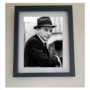 Puede incluir: Retrato enmarcado en blanco y negro de un hombre con sombrero fedora y traje. El retrato está en un marco gris oscuro con un passe-partout blanco. El hombre está mirando al espectador.