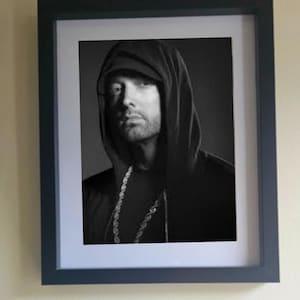 May include: Black and white framed portrait of a man wearing a hooded sweatshirt and a baseball cap. The man is also wearing a chain necklace. The portrait is in a black frame with a white mat.