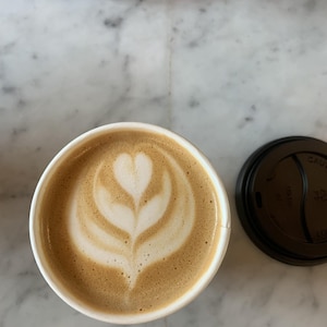 May include: A latte in a white paper cup with a heart-shaped design in the foam. The coffee is light brown, and the foam is white. A black lid sits next to the cup on a marble surface.
