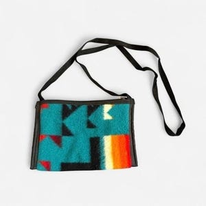 Puede incluir: Un bolso bandolera rectangular con una correa negra. El bolso presenta un patrón geométrico turquesa, negro, rojo y naranja. El bolso tiene una cremallera y un ribete negros.