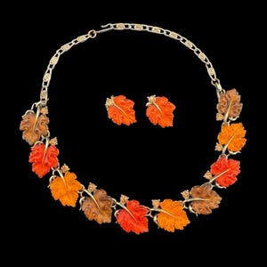May include: A gold-toned necklace and matching earrings set. The necklace and earrings feature leaf-shaped pendants in shades of orange, red, and brown, with small, sparkling accents. The jewelry set is displayed against a black background.